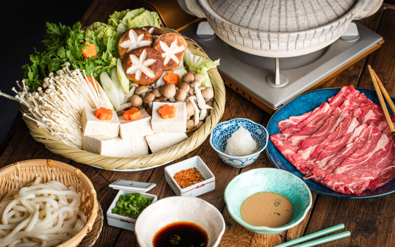A Japanese shabu-shabu hot pot setup with a donabe pot, thinly sliced beef, tofu, assorted mushrooms, vegetables, udon noodles, and dipping sauces arranged on a wooden table.